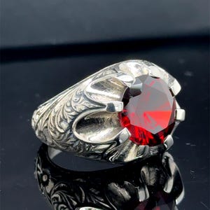 Könnte beinhalten: Ein silberner Ring mit einem ovalen, tiefroten Edelstein. Der Ring hat ein verziertes, graviertes Design auf dem Band. Der Edelstein ist in einer sicheren, zackenartigen Fassung gefasst, und der Ring ist hochreflektierend.