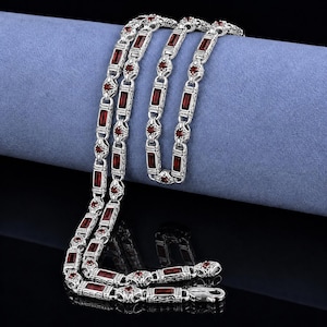 May include: Silver necklace featuring rectangular and round red gemstones. The chain is displayed on a blue surface, highlighting the intricate design and clasp. The gemstones are evenly spaced along the chain.