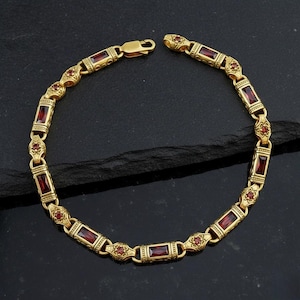 Może przedstawiać: Złoty bransoletka z prostokątnymi czerwonymi kamieniami szlachetnymi i ozdobnymi detalami. Bransoletka ma zapięcie i jest prezentowana na ciemnej powierzchni. Projekt zawiera wzory kwiatowe i geometryczne.