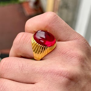 May include: A gold ring with a large, oval-shaped, red gemstone. The ring has a textured band.