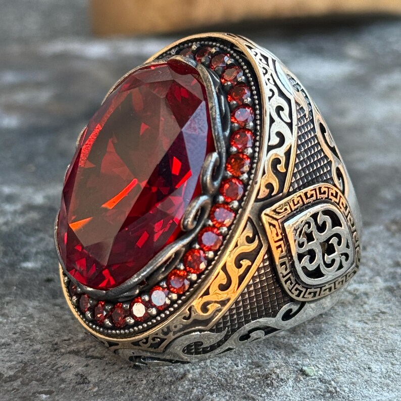 Puede incluir: Anillo plateado con una piedra preciosa roja ovalada y facetada. El anillo tiene un borde dorado alrededor de la piedra principal y est&aacute; adornado con piedras preciosas rojas m&aacute;s peque&ntilde;as. La banda del anillo presenta intrincados dise&ntilde;os grabados.