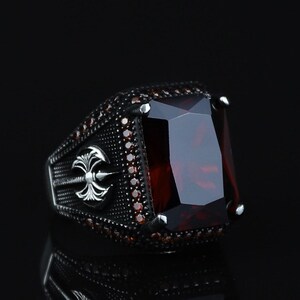 May include: A silver-toned ring with a large, rectangular, deep red gemstone as the centerpiece. The ring's band features intricate detailing and smaller red accent stones. The design includes a decorative emblem on the side, set against a dark background.