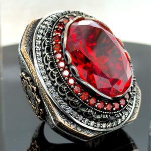 May include: A large, ornate ring featuring a faceted, oval-cut, deep red gemstone as the centerpiece. The ring has a silver-colored band with intricate detailing, including smaller red and clear gemstones. The design is elaborate and eye-catching.