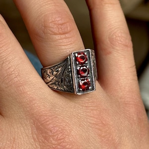 Könnte beinhalten: Ein silberner Ring mit einem verzierten Design und drei roten Edelsteinen, die in einer rechteckigen Form gefasst sind.