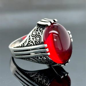 May include: A silver ring with an oval, deep red gemstone. The ring features intricate, swirling patterns on the band and a decorative top. The gemstone is smooth and polished, reflecting light.