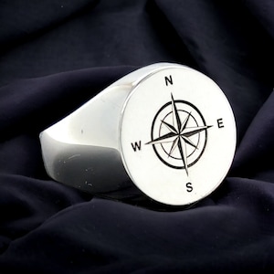May include: A silver signet ring with a compass design engraved on the top. The compass features the cardinal directions N, S, E, and W.