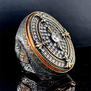 Puede incluir: Un anillo de plata con una gran cara de forma ovalada. La cara está decorada con un borde dorado y un diseño de cruz central con una piedra grande transparente. La cruz está rodeada de piedras transparentes más pequeñas.