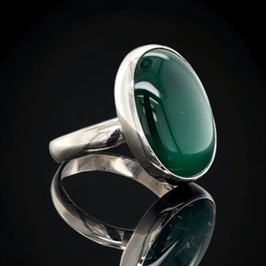 Puede incluir: Un anillo de plata con una gran piedra preciosa ovalada verde. El anillo está engastado en una banda simple y la piedra es lisa y pulida.