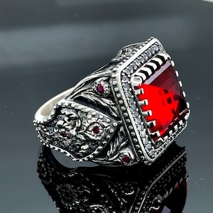 Könnte beinhalten: Ein silberner Ring mit einem großen roten Edelstein, der von kleineren weißen Edelsteinen umgeben ist. Der Ring hat aufwendige florale Designs auf der Band.