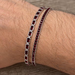 May include: Two silver-toned tennis bracelets worn together. One bracelet features a row of rectangular, deep red gemstones in a bezel setting, while the other bracelet displays a row of smaller, square-cut, deep red gemstones in a prong setting.