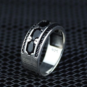 以下が含まれることがあります： テクスチャード加工されたバンドと黒い宝石が付いたシルバーリング。リングの中央には、3つの大きな楕円形の黒い石があり、その周りを小さな黒い石が囲んでいます。リングの内側には「925」の刻印があります。