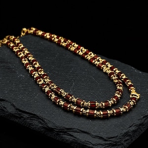 Könnte beinhalten: Eine Halskette mit einer Reihe zylindrischer, dunkelroter Perlen, die sich mit kleinen goldfarbenen Metallringen abwechseln. Die Kette ist in einem sich wiederholenden Muster aus diesen roten Segmenten und goldenen Verbindungsstücken gefertigt.