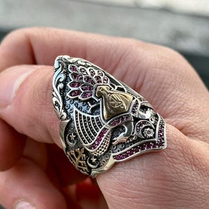Op de afbeelding: Een zilveren ring met een gedetailleerd ontwerp met een vogel met uitgespreide vleugels, een halve maan en sterren. De ring is versierd met rode edelstenen.