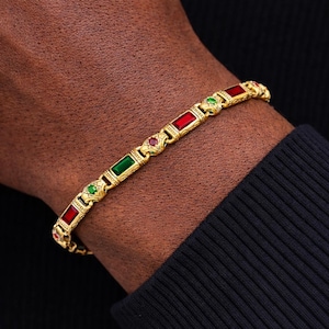 May include: Gold-toned bracelet featuring rectangular red and green gemstones. The bracelet has an ornate design with small, round gemstones between the rectangular stones. The bracelet is worn on a wrist against a dark background.