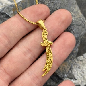 Op de afbeelding: Een goudkleurige hangerketting met een gebogen dolk ontwerp. De hanger heeft ingewikkelde details en hangt aan een fijne gouden ketting. De dolk heeft een gebogen lemmet met gegraveerde tekst.
