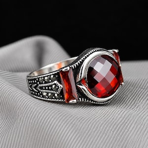Puede incluir: Un anillo de plata con una gran piedra roja redonda y facetada en el centro, flanqueada por dos piedras rojas rectangulares. El aro está decorado con intrincados trabajos en metal y pequeñas piedras oscuras incrustadas en los laterales.