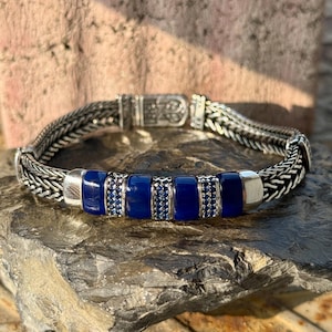 Peut inclure: Un bracelet en argent avec un motif de chaîne tissée. Le bracelet présente une rangée centrale de pierres rectangulaires bleu foncé, entrecoupées de petits accents bleus scintillants. Le fermoir est en argent et présente un design orné.