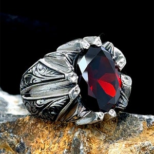 Może przedstawiać: Srebrny pierścionek z ozdobnym wzorem i dużym, owalnym, ciemnoczerwonym kamieniem szlachetnym.