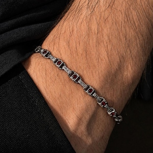 Könnte beinhalten: Silbernes Armband mit rechteckigen roten Edelsteinen. Das Armband wird an einem Handgelenk getragen und zeigt das aufwendige Design und den Kontrast zwischen dem silbernen Metall und den roten Steinen. Das Armband hat ein verziertes, vintage-inspiriertes Design.