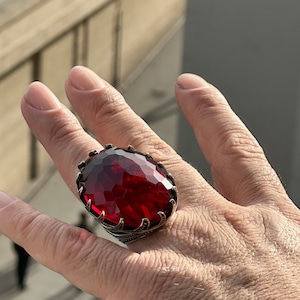 Op de afbeelding: Een zilveren ring met een grote, gefacetteerde, rode edelsteen. De ring wordt aan een vinger gedragen.