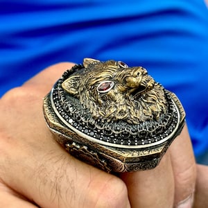 May include: A detailed, antique-style ring featuring a wolf's head. The ring is gold-toned with red gemstone eyes and black gemstone accents. The ring has an ornate, textured design.
