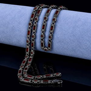 Op de afbeelding: Zilverkleurige ketting met rechthoekige rode edelstenen. De ketting heeft rechthoekige schakels met een rode steen in elk, afgewisseld met decoratieve bloemmotieven. De ketting wordt gepresenteerd op een lichtblauwe ondergrond.