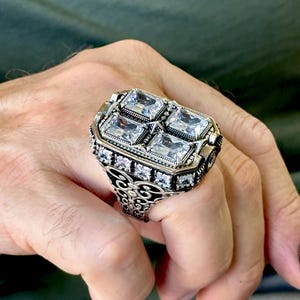 Puede incluir: Un anillo de plata con un diseño cuadrado que presenta cuatro piedras grandes transparentes. El anillo tiene detalles intrincados y una banda de filigrana.