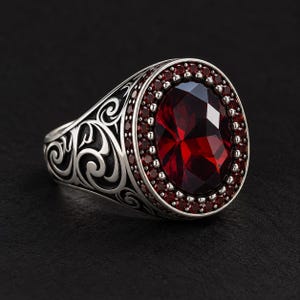 Op de afbeelding: Een zilveren ring met een grote, ovaal geslepen, dieprode edelsteen in het midden, omringd door een halo van kleinere rode steentjes. De ringband is gemaakt van geoxideerd zilver met gedetailleerde, verhoogde krulpatronen aan de zijkanten.