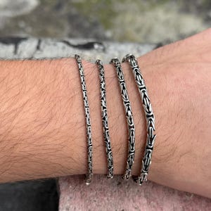 May include: Four silver chain bracelets of varying widths stacked on a person's wrist.