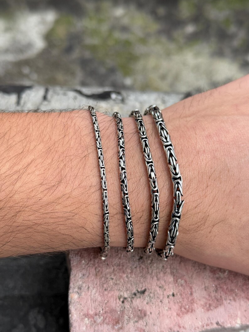 May include: Four silver chain bracelets of varying widths stacked on a person's wrist.