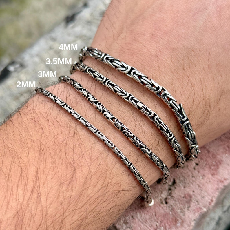 May include: Four silver chain bracelets of different widths, 2mm, 3mm, 3.5mm, and 4mm, are shown on a person's wrist. The bracelets are made of a silver chain with a box clasp.