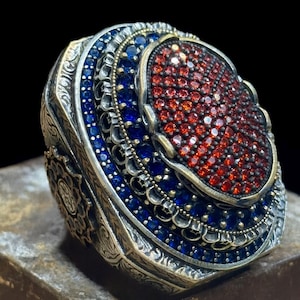 May include: Ornate silver ring with a large oval centerpiece. The center is covered in small red stones, surrounded by a layer of blue stones and intricate metalwork. The ring has a detailed, textured band.