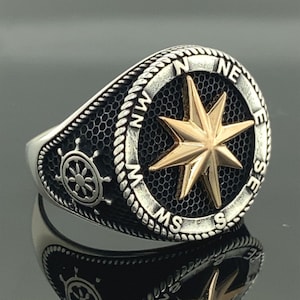 Puede incluir: Un anillo de plata con un diseño de brújula que presenta una estrella dorada y las letras N, E, S y W alrededor del borde.