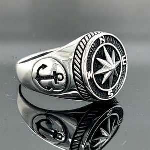 Può includere: Un anello d'argento con un designo di bussola e ancora. La bussola presenta un ago a forma di stella che punta a nord, sud, est e ovest. L'ancora è sul lato dell'anello.