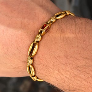 May include: A gold-toned chain bracelet featuring alternating smooth oval links and textured links with intricate patterns. The bracelet includes a single rectangular link set with a deep red gemstone, creating a distinct focal point.