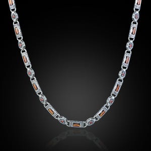 May include: Silver-toned necklace with rectangular amber-colored stones and small red accents. The chain links are detailed with floral and geometric patterns. The necklace is displayed against a dark background, highlighting its intricate design.