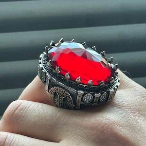 Könnte beinhalten: Ein silberner Ring mit einem großen, roten, facettierten Edelstein, der in einem kronenartigen Design gefasst ist. Der Ring hat komplizierte Details und eine silberne Band.
