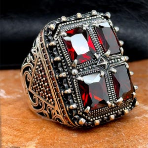 Könnte beinhalten: Ein silberner Ring mit einem großen, quadratischen roten Edelstein, der in der Mitte eingesetzt ist. Der Ring ist mit komplizierten Details und kleineren roten Edelsteinen um das Band verziert.