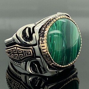 Puede incluir: Un anillo de plata con una piedra de malaquita verde engastada en un bisel de oro. El anillo tiene un diseño detallado con un patrón de llave griega.