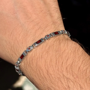 May include: Silver bracelet with rectangular and round red gemstones. The bracelet is worn on a person's wrist. The bracelet has a detailed design with small, round red gemstones between the larger rectangular stones.