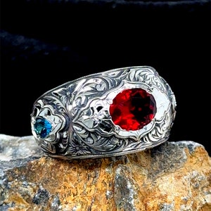 May include: A silver ring with an ornate design featuring a large red gemstone and two smaller blue gemstones.