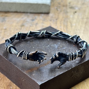 May include: A silver bracelet with two hands reaching out to each other. The bracelet is made of twisted wire and has a dark oxidized finish.