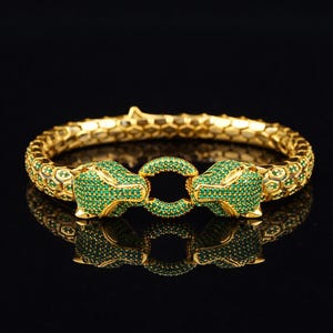 May include: A gold-toned bracelet featuring two panther heads encrusted with green gemstones. The bracelet has a textured, snake-like design and a circular clasp. The jewelry is displayed on a reflective black surface.