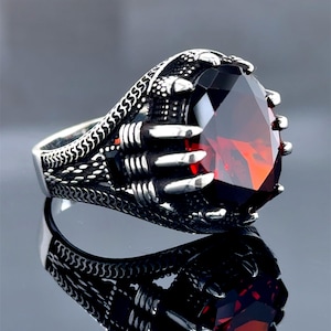 Peut inclure: Une bague en argent avec une grande pierre rouge sertie dans un motif en forme de griffe. La bande de la bague est ornée d'un motif texturé.