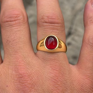 Op de afbeelding: Een goudkleurige ring met een gladde, ovaalvormige, dieprode cabochon edelsteen in het midden. De band heeft een gepolijste, reflecterende afwerking met een solide, afgerond ontwerp dat iets taps toeloopt naar de achterkant van de vinger.