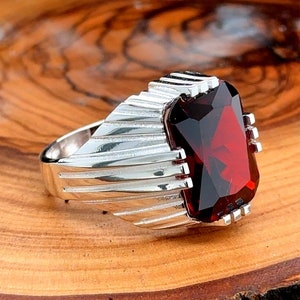 Może przedstawiać: Srebrny pierścionek z dużym, ciemnoczerwonym kamieniem szlachetnym. Pierścionek ma fakturowany pasek z rowkowanym wzorem.