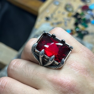 Op de afbeelding: Een zilveren ring met een grote, rode, gefacetteerde edelsteen in een klauwsetting. De ring is ontworpen om eruit te zien als een set klauwen.