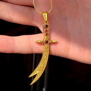 Op de afbeelding: Goudkleurige hangerketting met een gebogen zwaardontwerp. De hanger heeft rode edelsteenaccenten en gedetailleerde patronen. De ketting hangt aan een fijne gouden ketting.