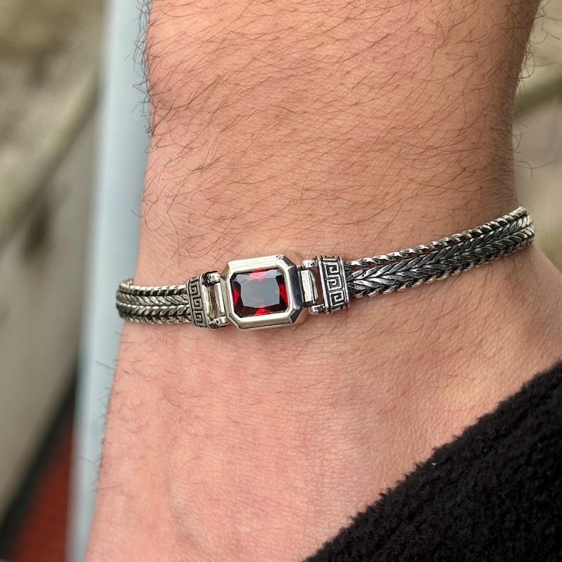May include: A silver bracelet with a rectangular, deep red gemstone at the center. The bracelet features a braided chain design with decorative geometric patterns on either side of the gemstone. The bracelet is worn on a wrist.
