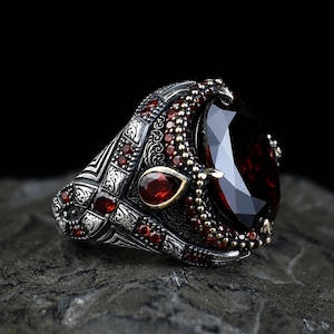 May include: A silver ring featuring a large, oval-cut dark red gemstone as the centrepiece. The band is decorated with intricate metal engravings and accented with numerous small, round, and pear-shaped red stones set along the sides and frame.
